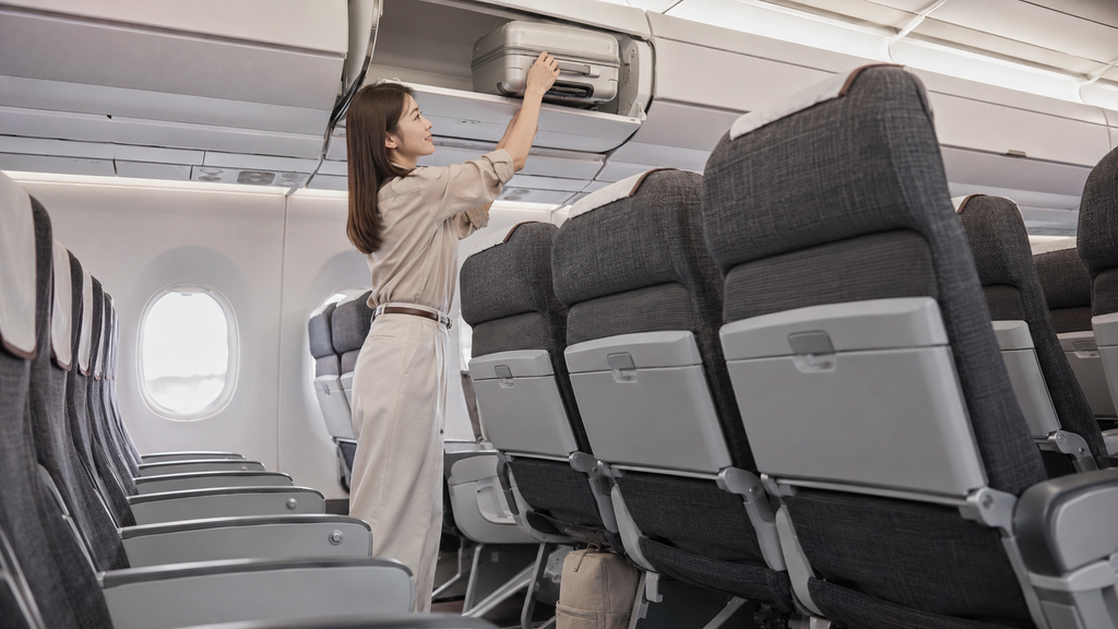 機内持ち込みスーツケースを収納棚へ入れる日本人女性と前席下のリュック